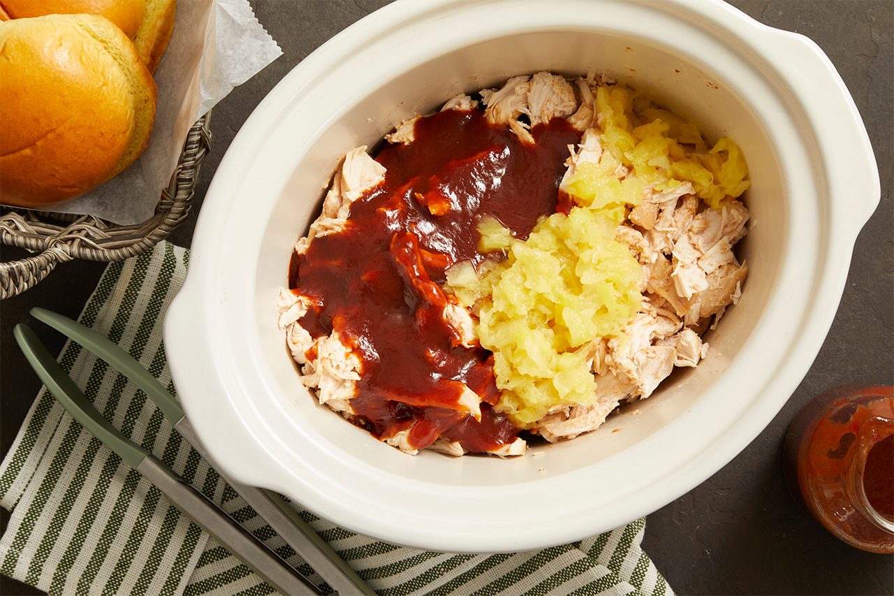 Shredded chicken in a slow cooker topped with barbecue sauce and crushed pineapple, next to a basket of sandwich buns and a green striped napkin with utensils.