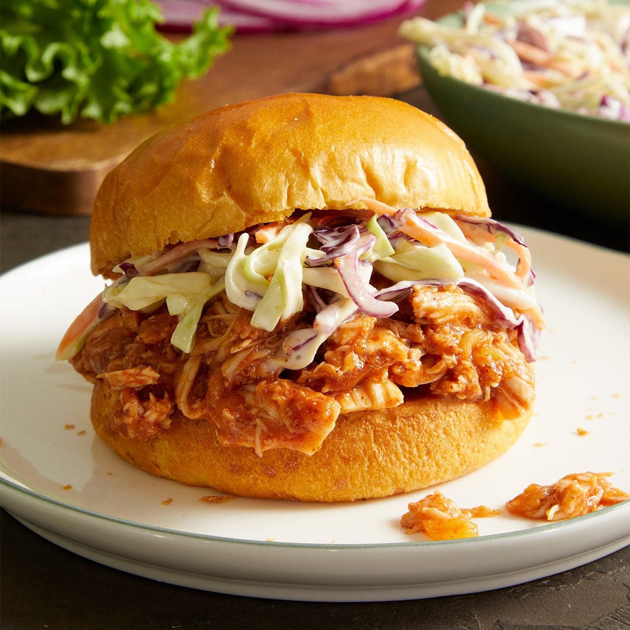 Pulled pork sandwich topped with creamy coleslaw, served on a white plate. A bowl of coleslaw and fresh lettuce are in the background.