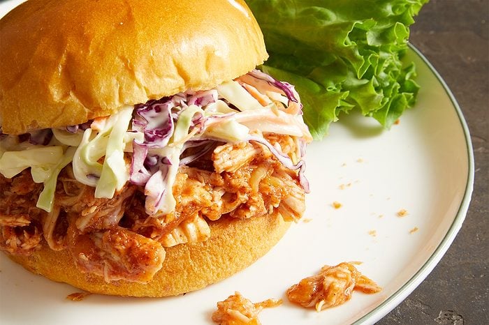 Pulled chicken sandwich with creamy coleslaw on a bun, served on a white plate with a leaf of green lettuce on the side.