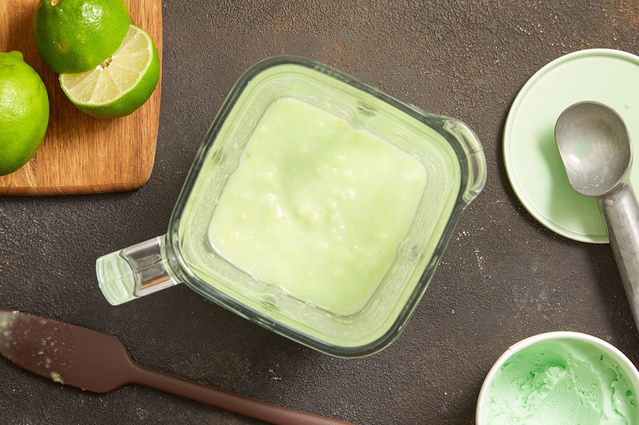 Overhead horizontal image of a light green mixture in a blender, flanked by limes on a wooden cutting board to the left. A spatula rests below the blender, while an ice cream scoop on a light green plate and a small container of green ice cream are visible on the right.