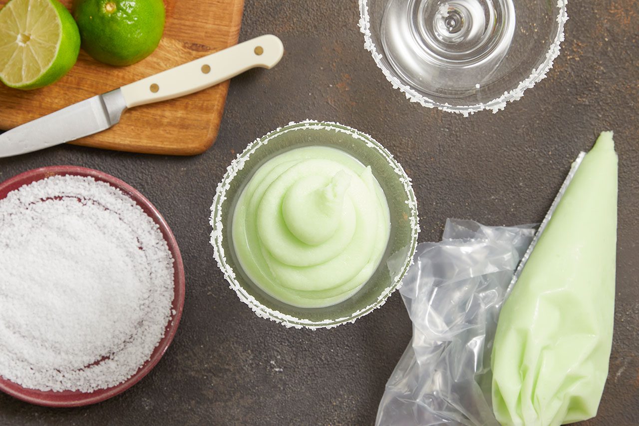 Overhead horizontal image of a margarita glass being filled with a light green frozen drink from a piping bag. Visible are whole and halved limes on a wooden cutting board with a knife, a bowl of salt, and another empty salted glass, all on a dark background.
