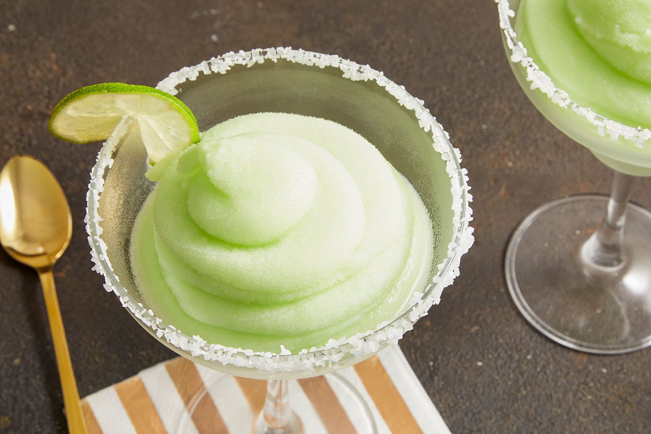 Overhead horizontal image of two frosted margarita glasses filled with a vibrant light green frozen drink. The rims of the glasses are coated with salt, and the foreground glass is garnished with a fresh lime wedge. A gold spoon and a striped napkin are partially visible on the dark background.