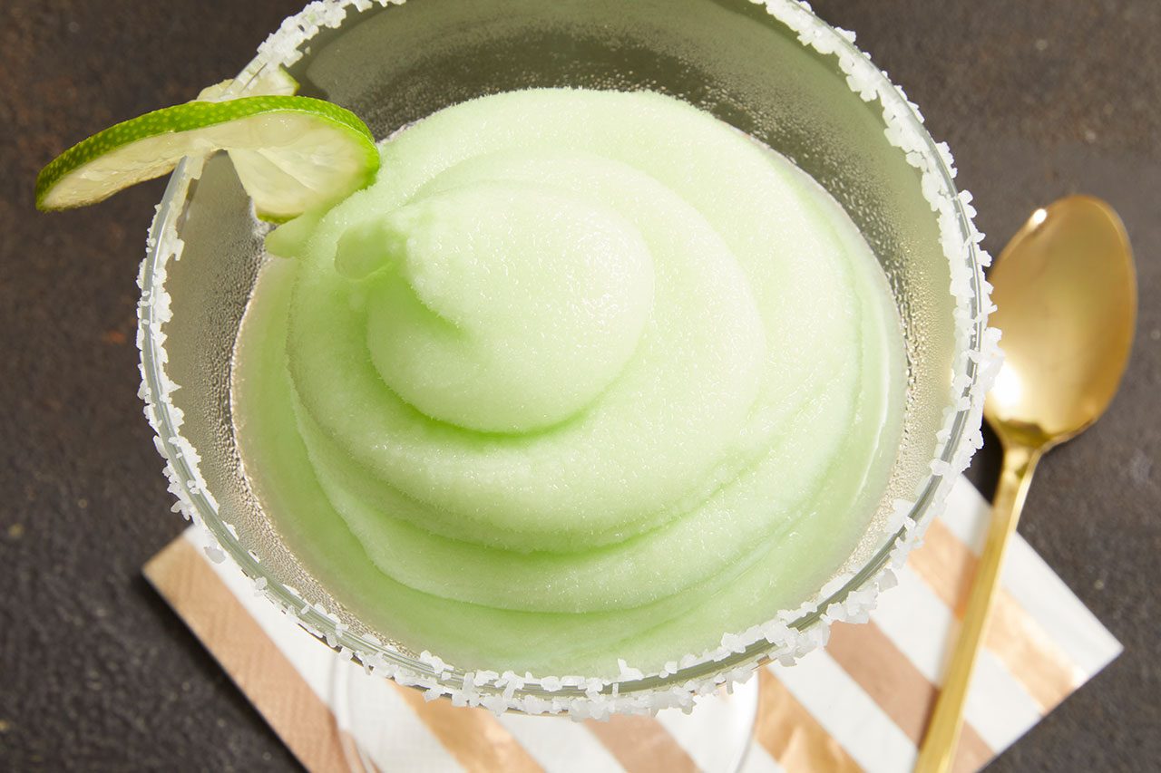 Overhead horizontal image of a light green frozen drink in a salt-rimmed margarita glass. A lime slice garnishes the side of the glass, which sits on a striped napkin next to a gold spoon.