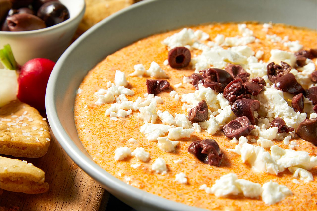 Eye Level horizontal image of a creamy orange dip, garnished with crumbled feta cheese and chopped Kalamata olives, served in a gray bowl. The dip is accompanied by crackers, a radish, and an onion on a wooden surface, with a small bowl of whole olives visible in the background.