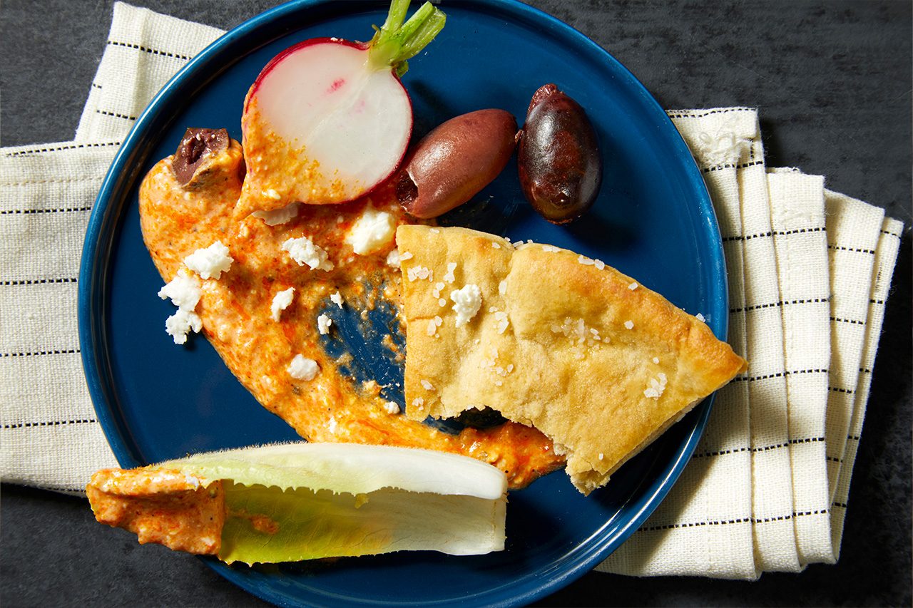 Overhead horizontal image of a blue plate holding a creamy orange dip topped with crumbled feta cheese. The plate also features a piece of pita bread, a halved radish, two dark olives, and an endive leaf, all resting on a dark surface next to a striped napkin.