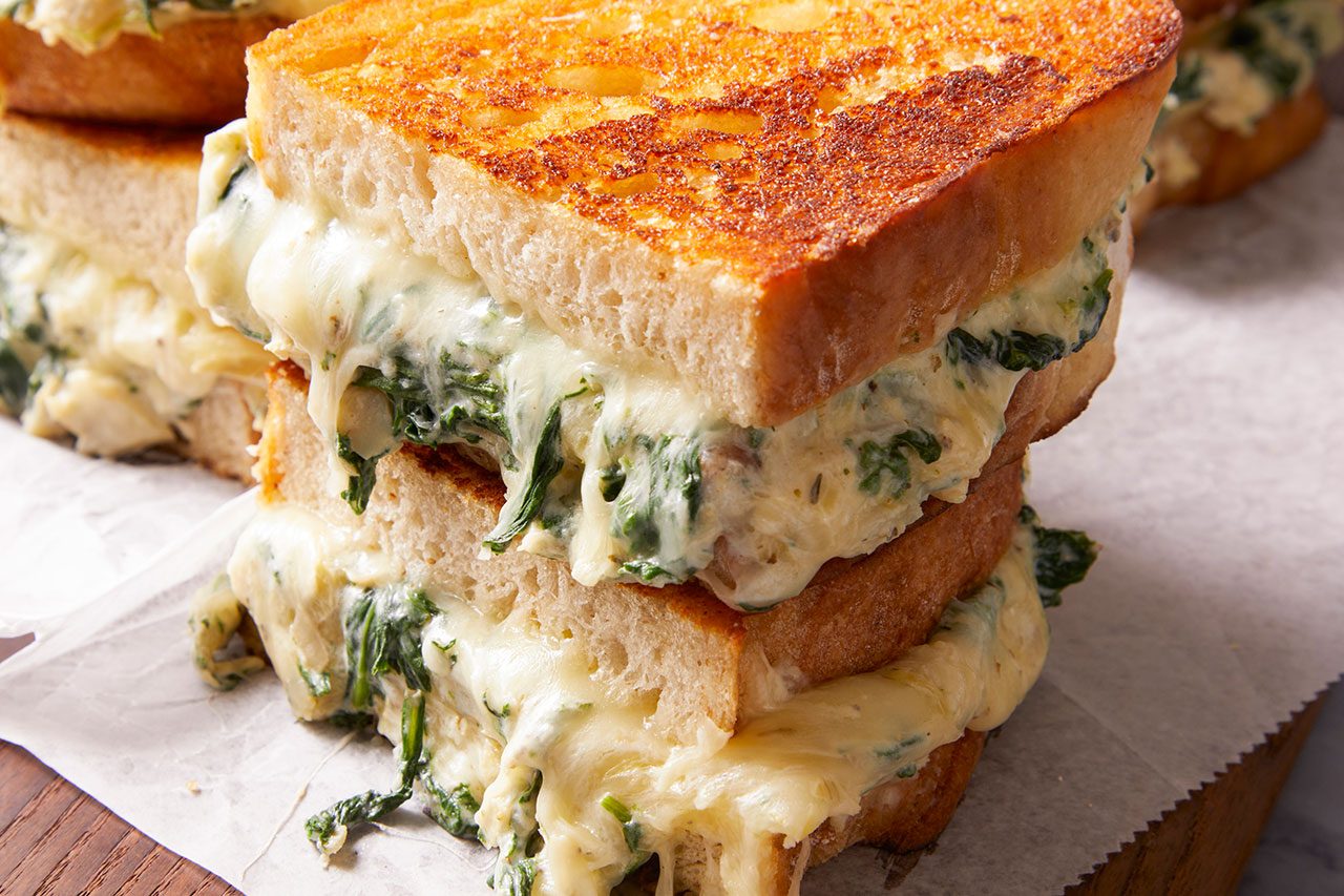 Eye Level horizontal image of two halves of a stacked, gooey grilled cheese sandwich. The golden-brown toasted bread is filled with a rich, melted cheese and spinach mixture, which oozes temptingly from the sides. The sandwiches are presented on a piece of white parchment paper.