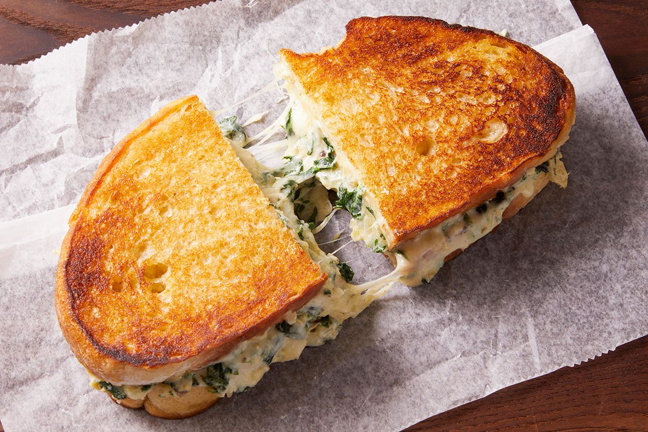 Overhead horizontal image of a golden brown grilled cheese sandwich, cut in half, revealing a creamy filling of melted cheese and spinach with visible cheese pulls. It rests on a crinkled piece of white parchment paper.