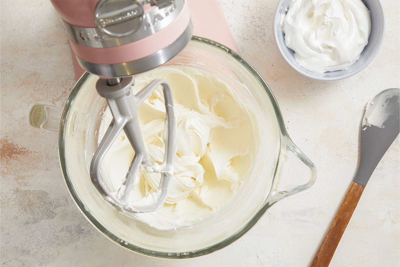 Overhead horizontal image of a pink KitchenAid stand mixer with its paddle attachment mixing a white, creamy frosting or whipped cream in a clear glass bowl. A small gray bowl filled with more of the creamy substance and a spatula with residue are also visible on the light-colored counter surface.