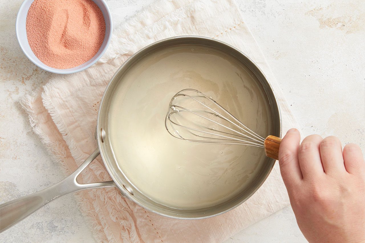 Overhead horizontal image of a hand whisking a light-colored liquid in a stainless steel saucepan. A small white bowl filled with pink-orange colored powder is visible in the upper left corner, resting on a light-colored cloth.