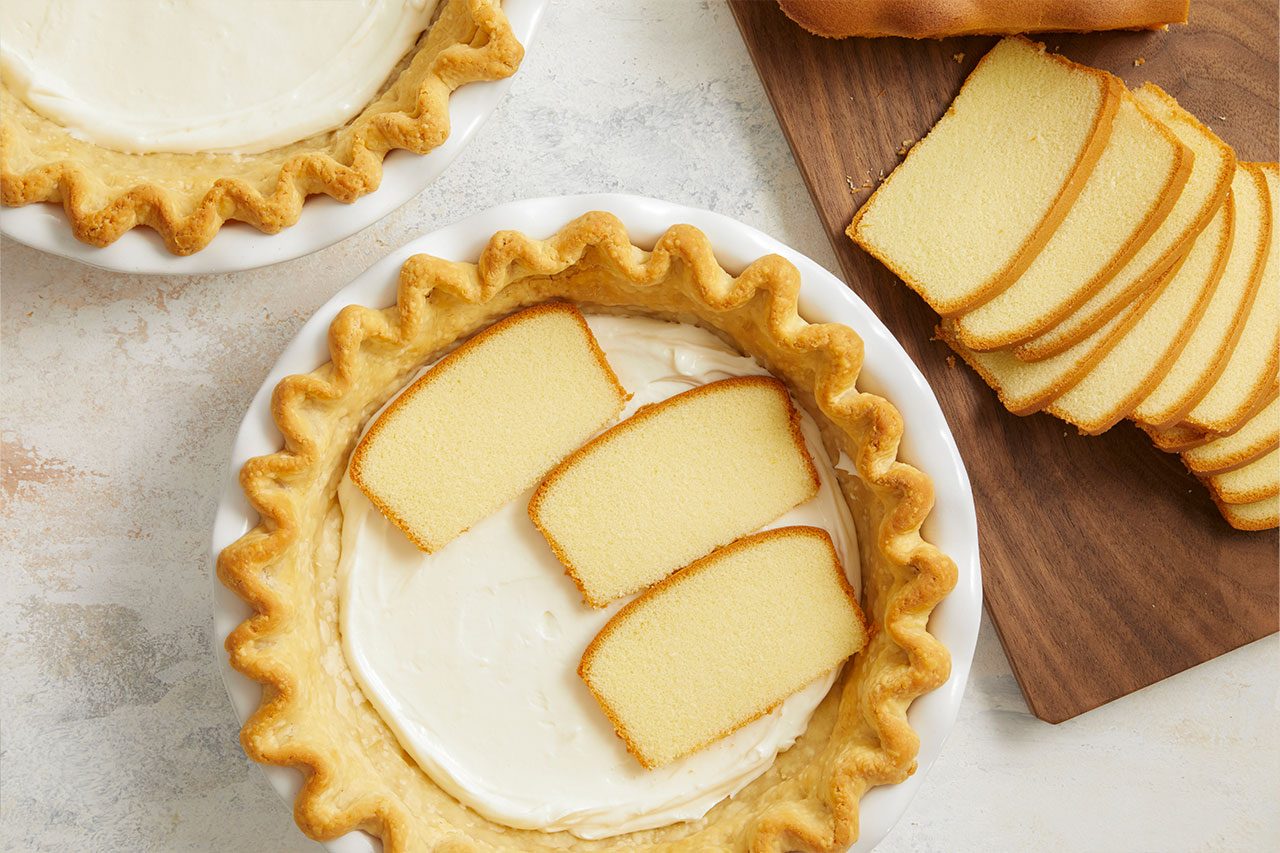Overhead horizontal image of a pie dish filled with a creamy white layer, topped with three slices of golden pound cake. In the background, another pie dish with the same creamy layer is partially visible, while a wooden cutting board to the right holds several fanned-out slices of pound cake next to a partial loaf.