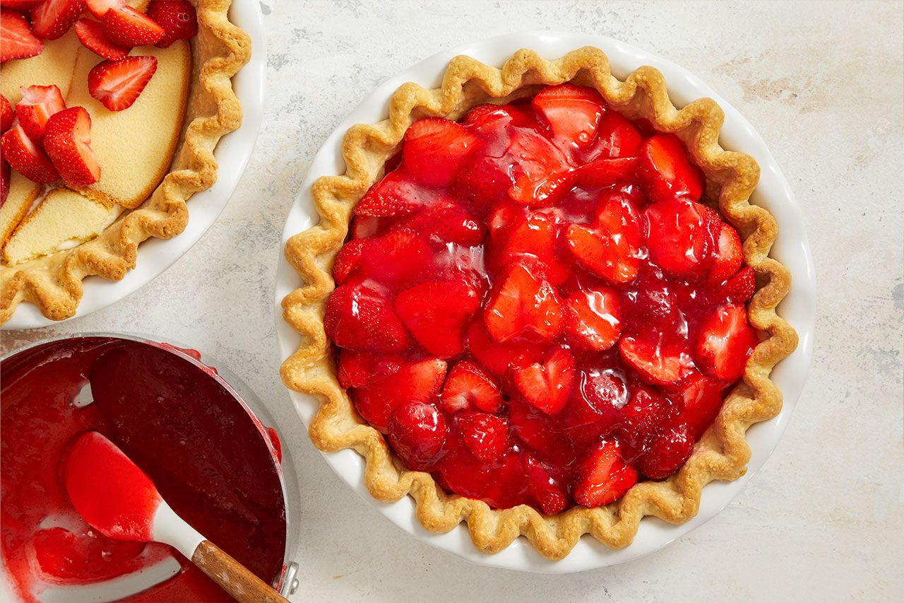 Overhead horizontal image of a freshly made strawberry pie in a white fluted pie dish, brimming with glazed fresh strawberries. To its left, a partially visible pie crust holds slices of cake topped with fresh strawberries, and a small saucepan with strawberry glaze and a spatula.