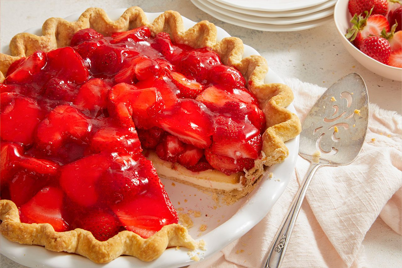 Overhead horizontal image of a glossy strawberry pie in a white pie dish, with one slice removed revealing a creamy white filling beneath the fruit. A pie server rests on a light pink napkin beside the pie, while a bowl of fresh strawberries and a stack of plates are visible in the background.