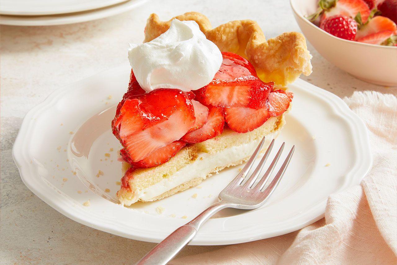 Eye Level horizontal image of a delicious slice of strawberry pie. The pie slice features a flaky crust, a creamy white filling, and is generously topped with fresh sliced strawberries and a dollop of whipped cream, served on a white plate with a fork.