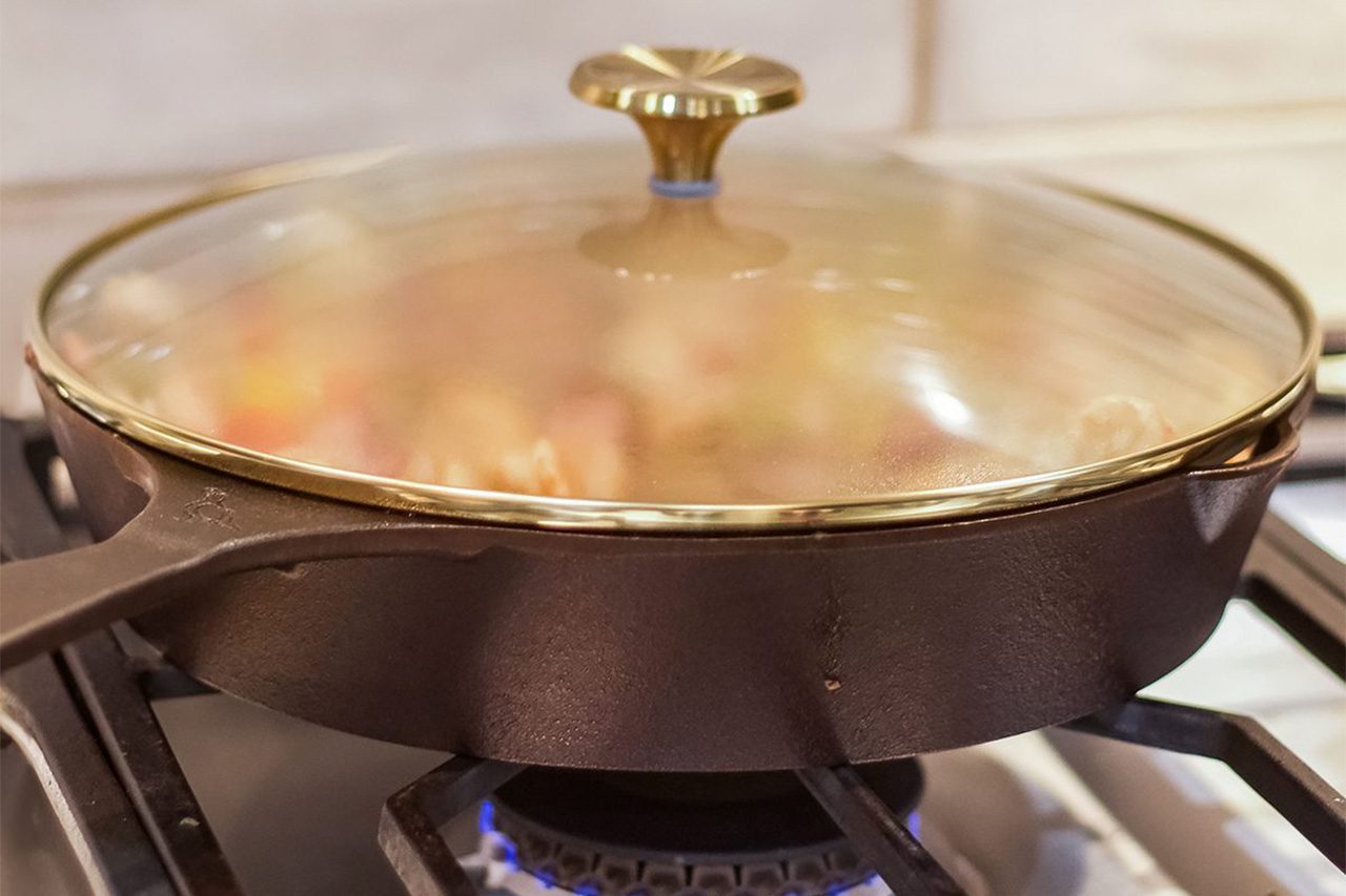 A black skillet with a glass lid sits on a gas stove burner with a blue flame visible underneath. The contents inside appear to be cooking and are slightly blurred under the lid.