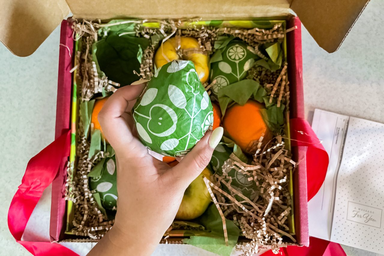 A hand holds a green, patterned fruit, with more assorted fruits and decorative packing material inside an open gift box. Red ribbon and a note are visible beside the box.