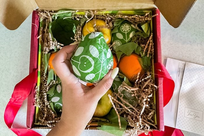 A hand holds a green, patterned fruit, with more assorted fruits and decorative packing material inside an open gift box. Red ribbon and a note are visible beside the box.