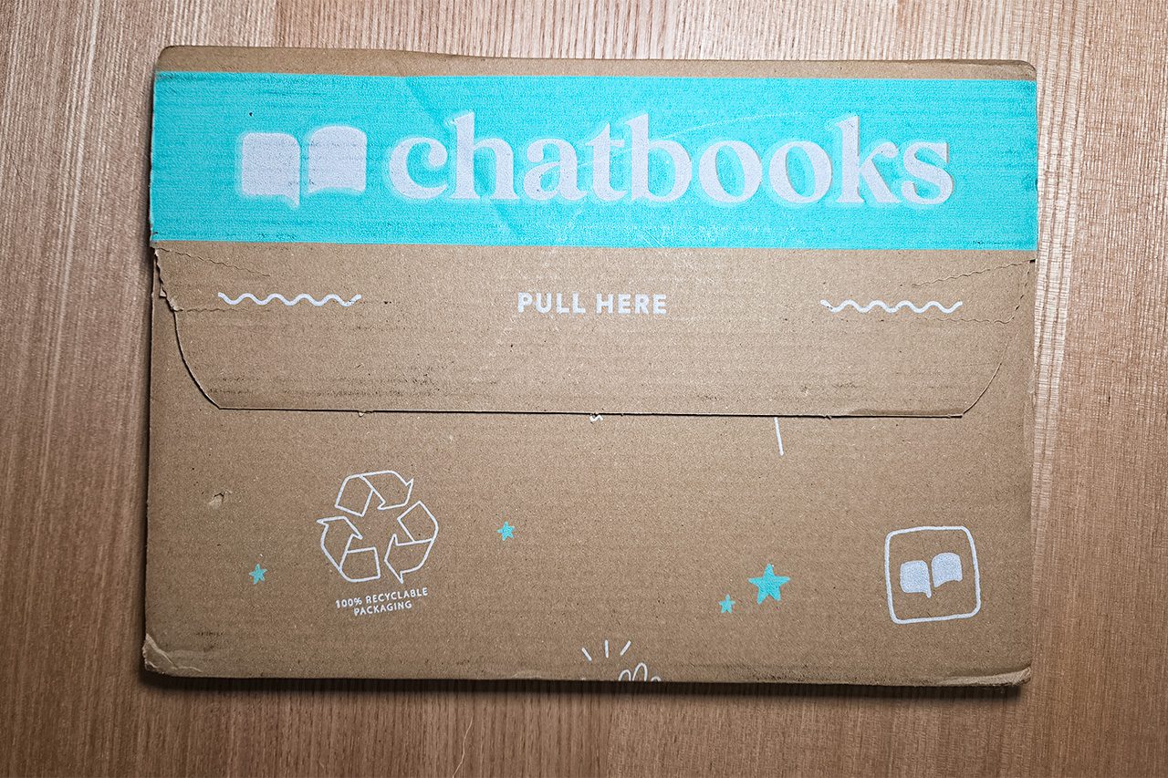 A cardboard envelope with a teal flap labeled “chatbooks,” featuring a recycle symbol and the instruction “PULL HERE” on a wooden surface.