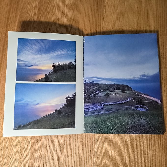 An open book on a wooden surface displays landscape photos: two sunset images on the left page and a wide coastal view with grassy hills on the right page.