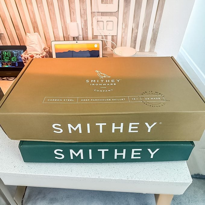 Two Smithey branded boxes, one brown and one green, stacked on a white table with various electronics and decor in the background.