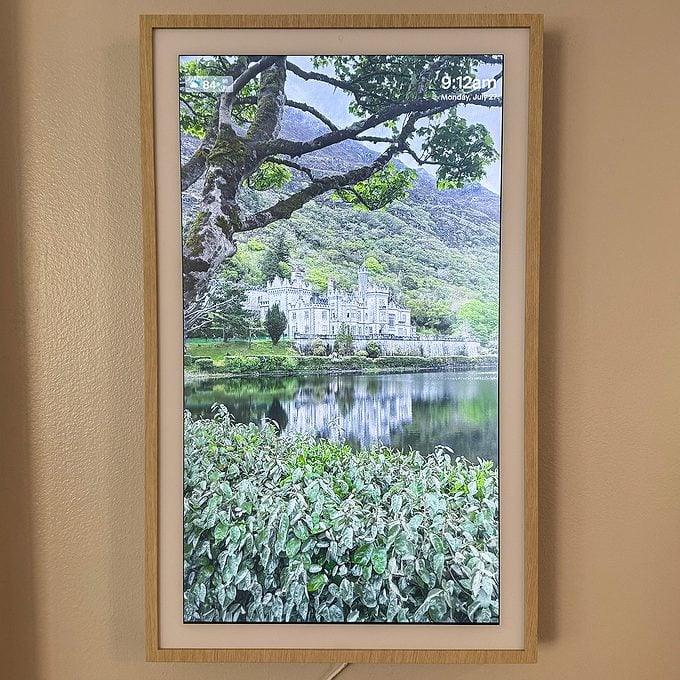 A digital photo frame displays a scenic landscape featuring a lake, dense green foliage, a large tree, and a castle-like building in the background under a cloudy sky.