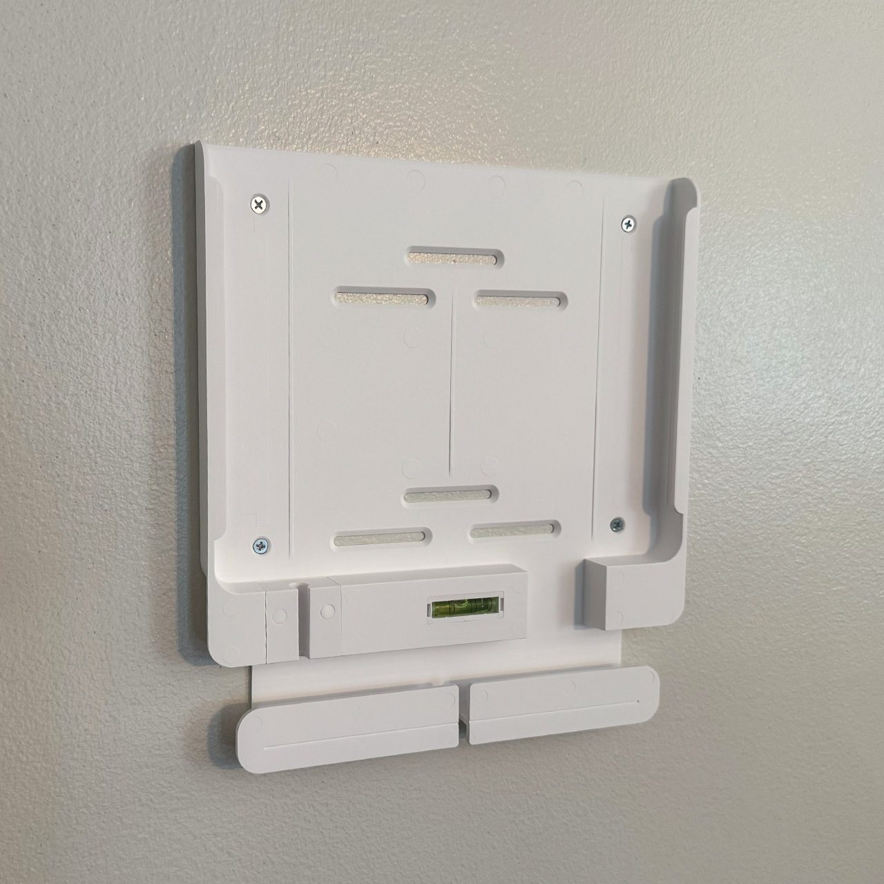 A white TV wall mount bracket with a built-in level is attached to a beige wall. It is square-shaped with slotted openings and four visible screws.