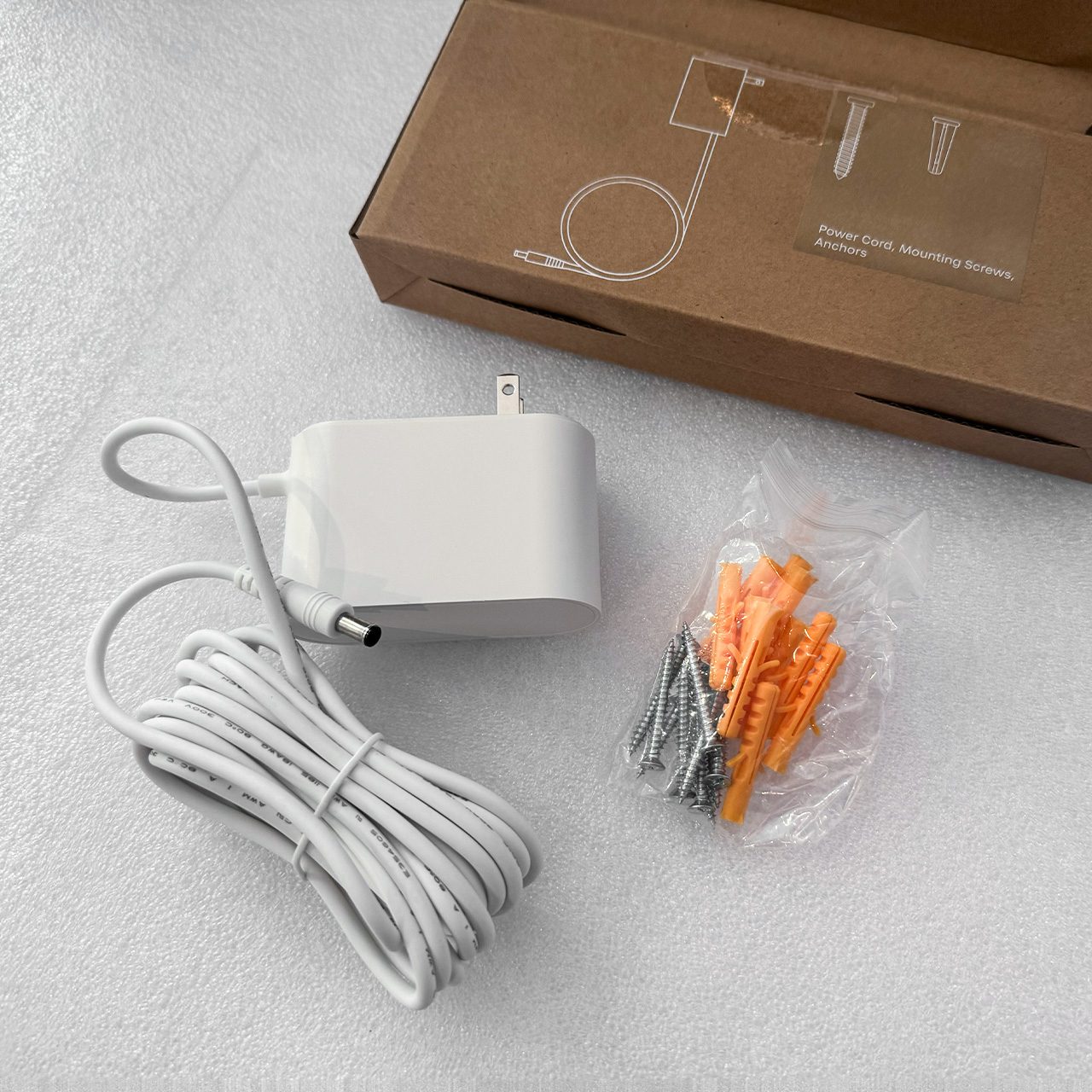 A white power adapter with a long cord, a small bag of screws and wall anchors, and a brown cardboard box with installation instructions printed on it are on a white surface.