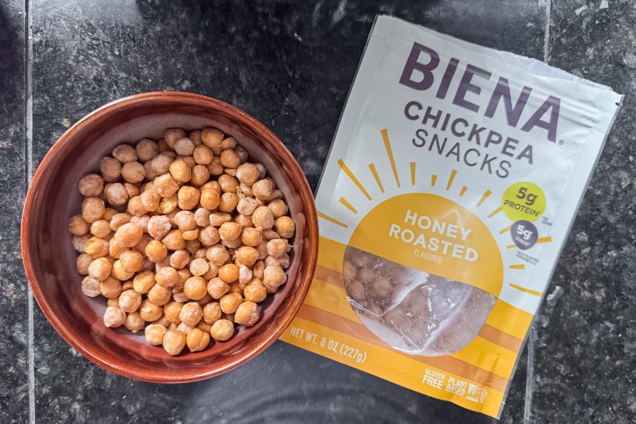 A bowl of honey roasted chickpeas sits next to a partially opened bag of Biena Chickpea Snacks on a dark countertop.