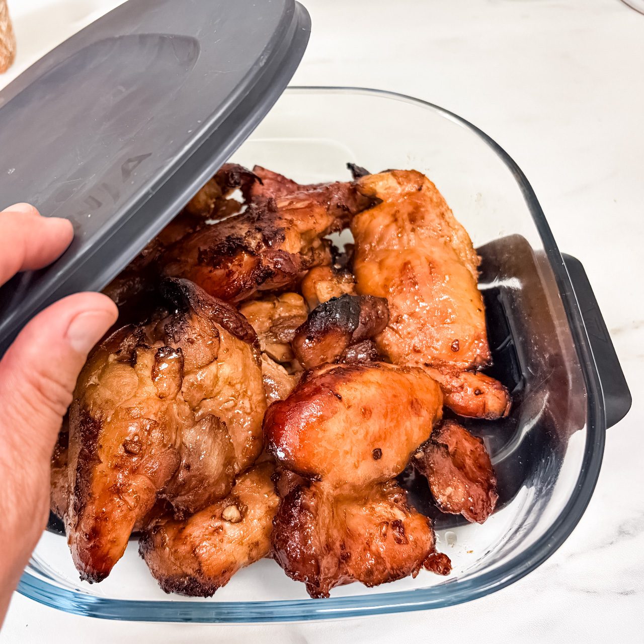 A hand lifts the lid off a glass container filled with cooked, browned chicken pieces on a white countertop.