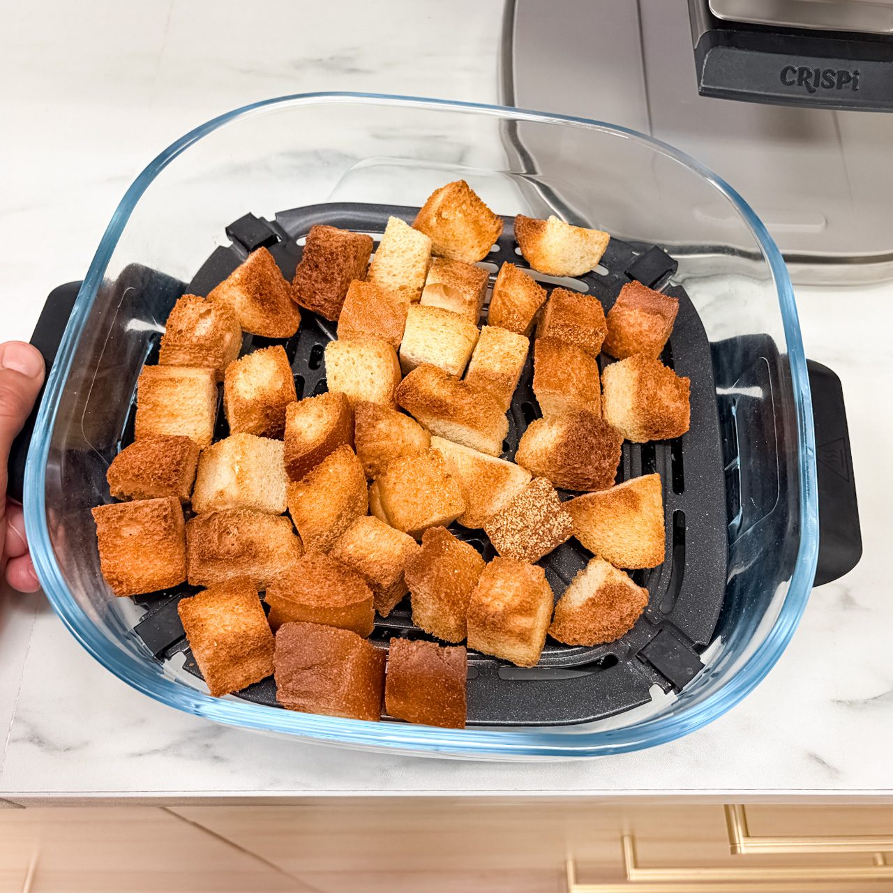 A glass dish filled with evenly browned croutons sits on a black tray, resting on a light kitchen countertop near an appliance.