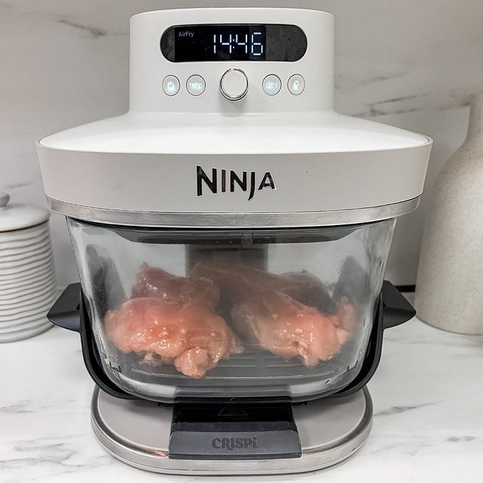 Raw chicken breasts inside a Ninja air fryer oven, with the digital display showing a time of 14:46. The appliance is on a kitchen counter next to stacked plates and a white container.