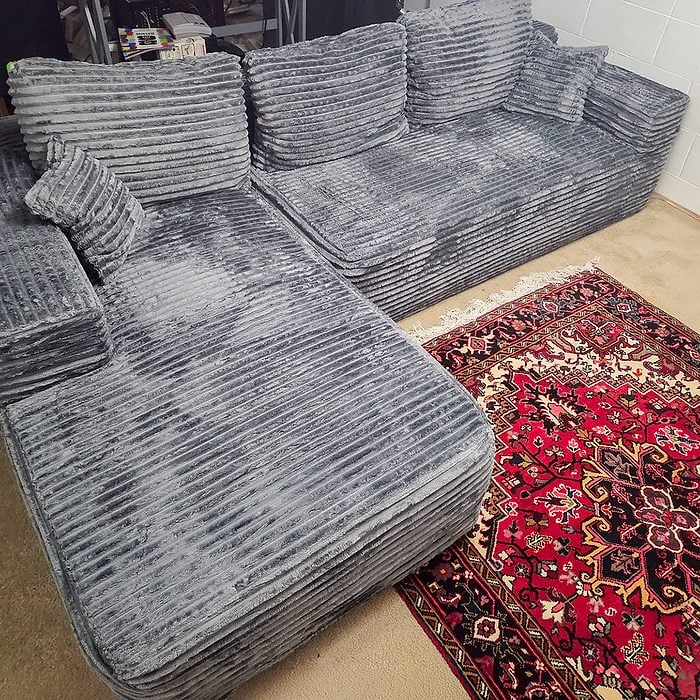 A gray, corduroy sectional sofa with matching cushions sits next to a red patterned rug on a light-colored carpeted floor.