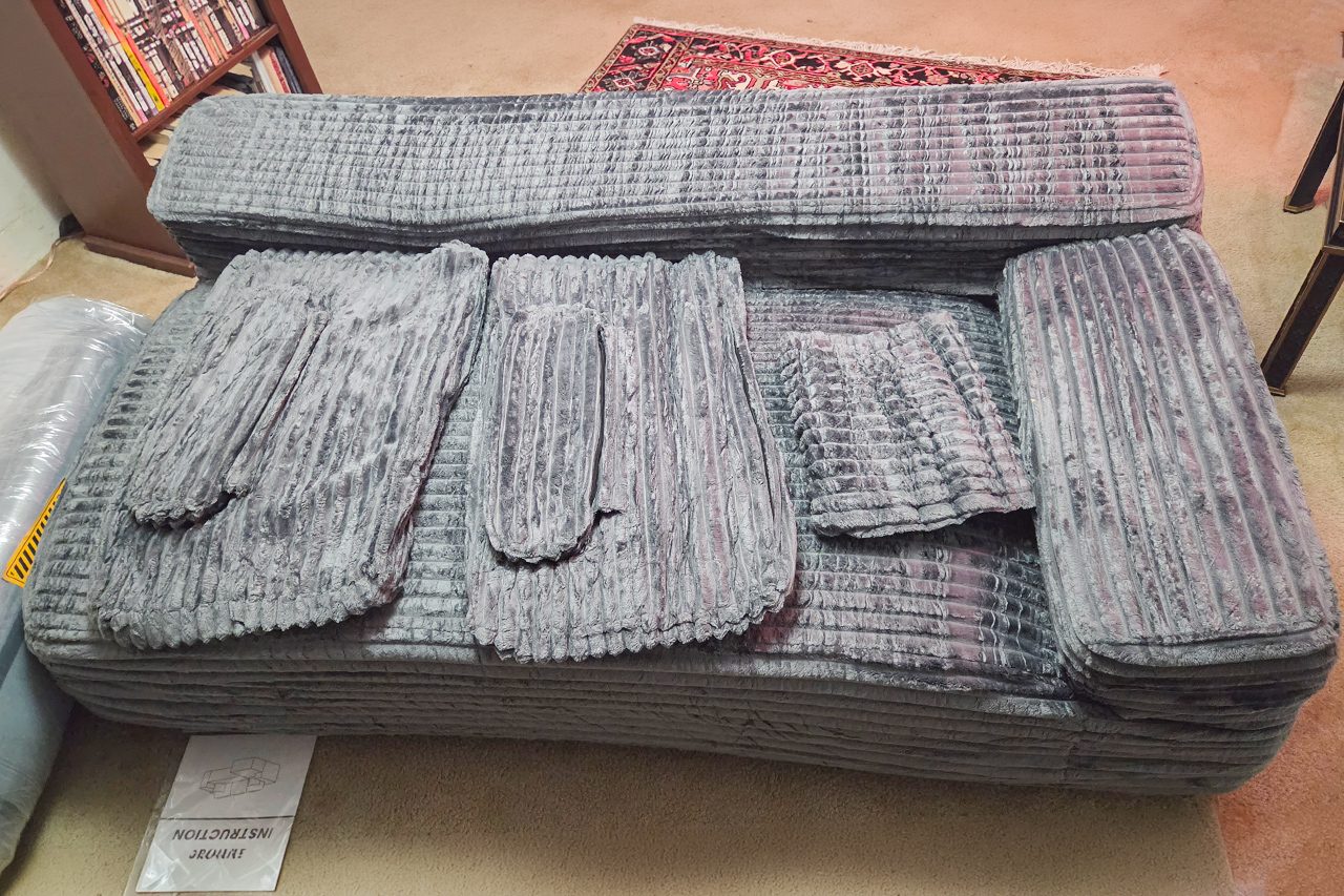 A gray corduroy sofa with its cushions removed and placed haphazardly on top, exposing the seat. A rug and shelves are partially visible in the background.