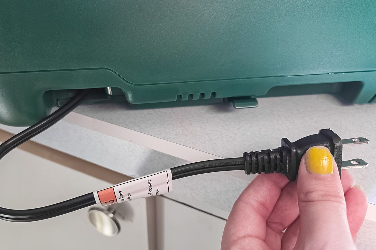A hand with yellow nail polish holds a power plug near an unplugged green electronic device sitting on a light countertop.