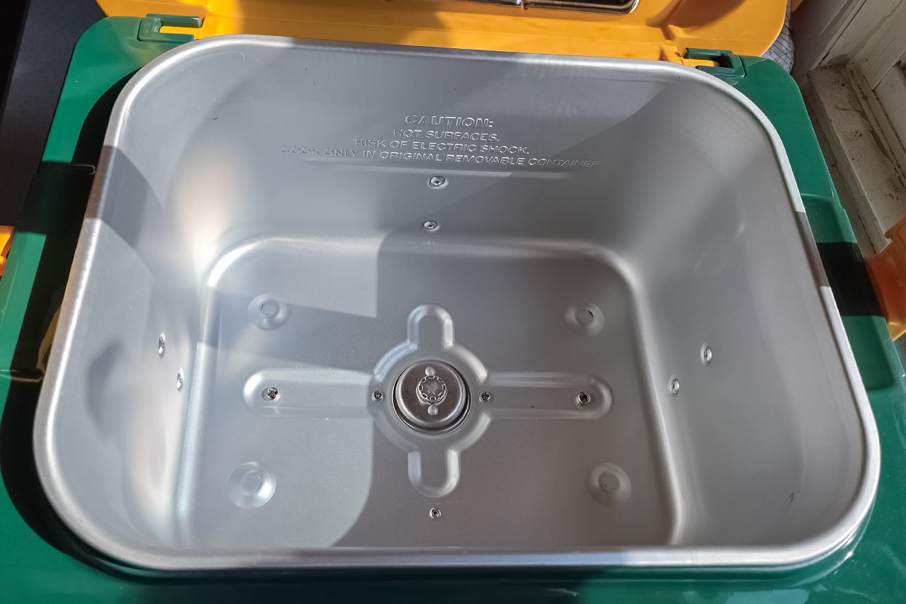 Open, empty, rectangular metal container with rounded corners and embossed text on the back wall; set inside a green and yellow exterior with a hinged lid.