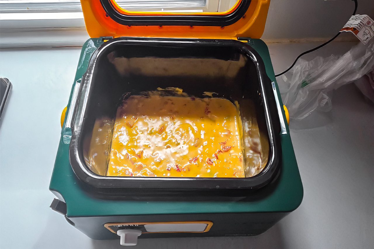 A portable electric cooker with its lid open shows an egg mixture with visible diced ingredients, possibly being prepared as an omelet, on a kitchen countertop.