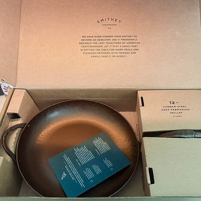 An open box contains a Smithey cast iron skillet with a handle, accompanied by an informational card and protective packaging.