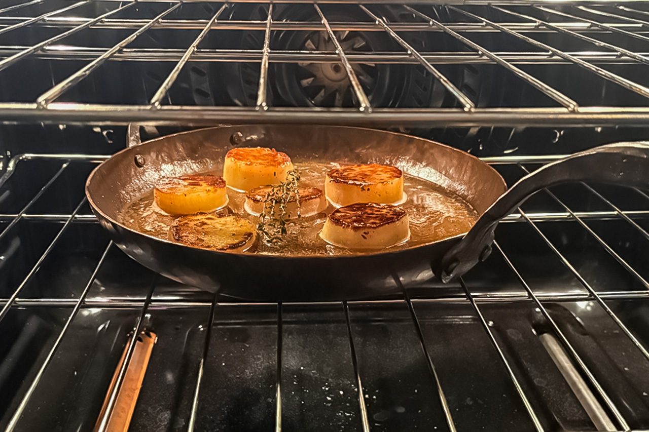A cast iron skillet with five seared scallops is placed on an oven rack, with the oven door open slightly, and the scallops are browning on top.