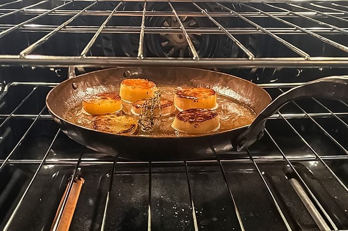 A cast iron skillet with five seared scallops is placed on an oven rack, with the oven door open slightly, and the scallops are browning on top.