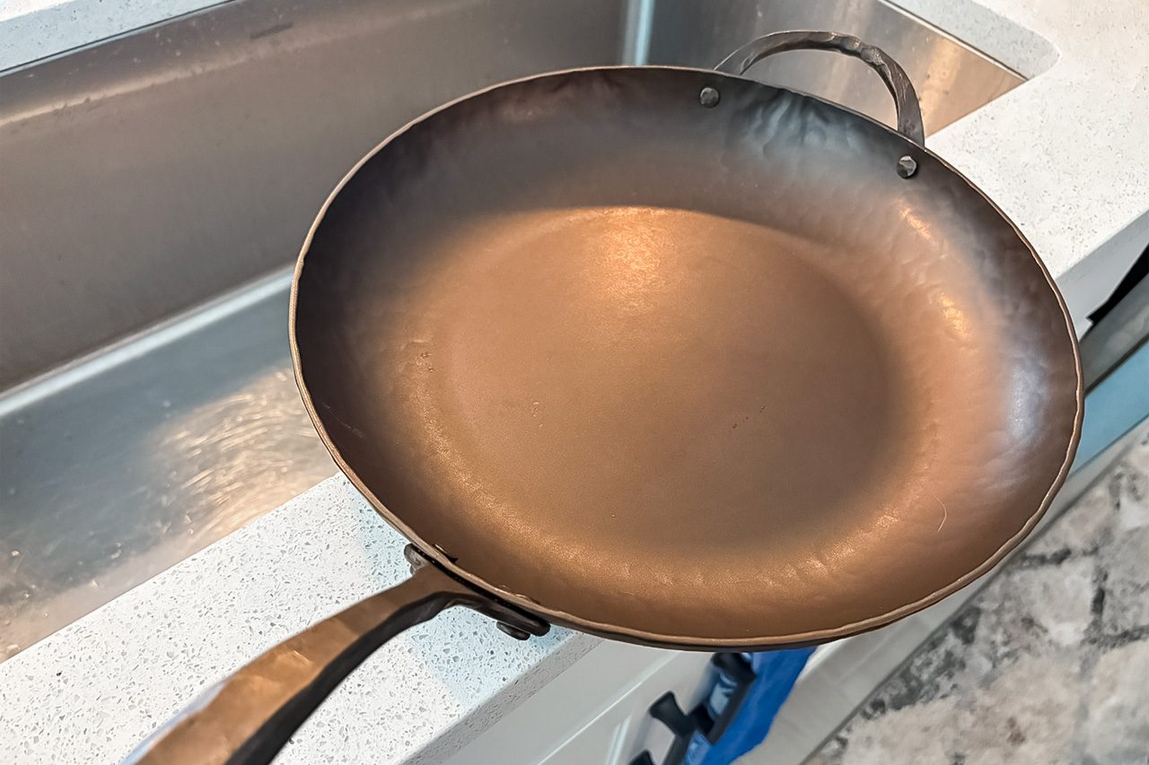 A clean, round metal frying pan with handles sits next to a kitchen sink on a countertop.