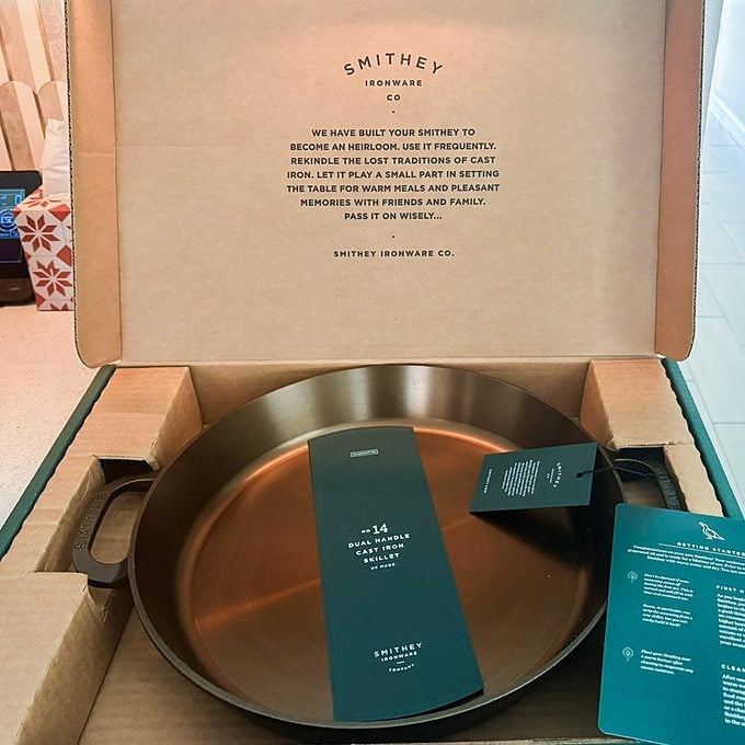 An open cardboard box displays a Smithey cast iron skillet with tags attached, resting in protective packaging. Text is printed inside the box lid, and a green card with product information is included.