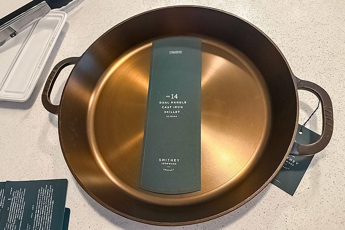 A round, bronze-colored cast iron pan with two handles sits on a countertop. A green product label with text is placed inside the pan.