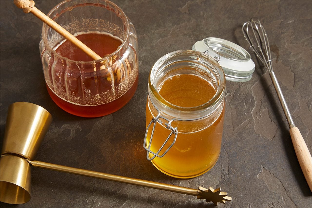 Overhead shot of honey and a golden honey mixture prepared in glass jars, with a honey dipper, whisk, and measuring cup arranged nearby on a dark stone surface.
