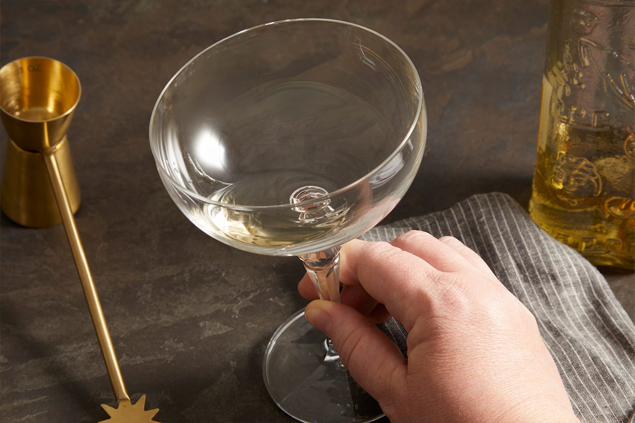 Close-up shot of an empty coupe glass being held by the stem, with a gold jigger, honey dipper, and bottle arranged nearby on a dark stone surface.
