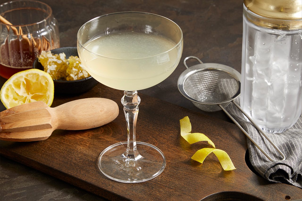 3/4 angle shot of a Tequila Honey Bee cocktail served in a coupe glass, featuring a pale golden, citrusy drink garnished with a lemon peel. Fresh lemon, honey, a strainer, and cocktail-making tools are arranged nearby on a dark wooden surface.