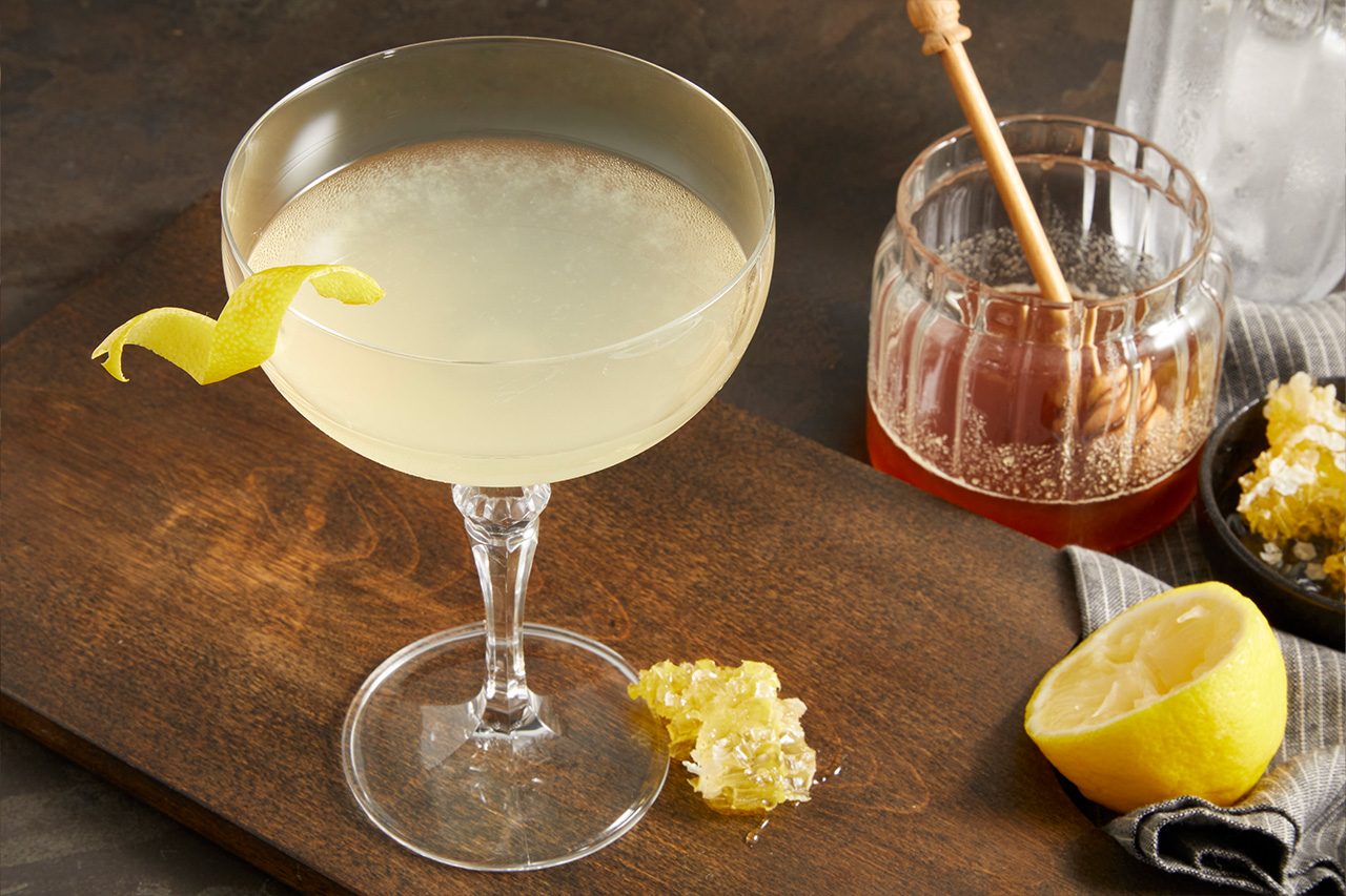 3/4 angle shot of a Tequila Honey Bee cocktail served in a coupe glass, featuring a pale golden drink garnished with a curled lemon peel. A honey jar with a dipper, fresh lemon half, and honeycomb are arranged nearby on a dark wooden surface.