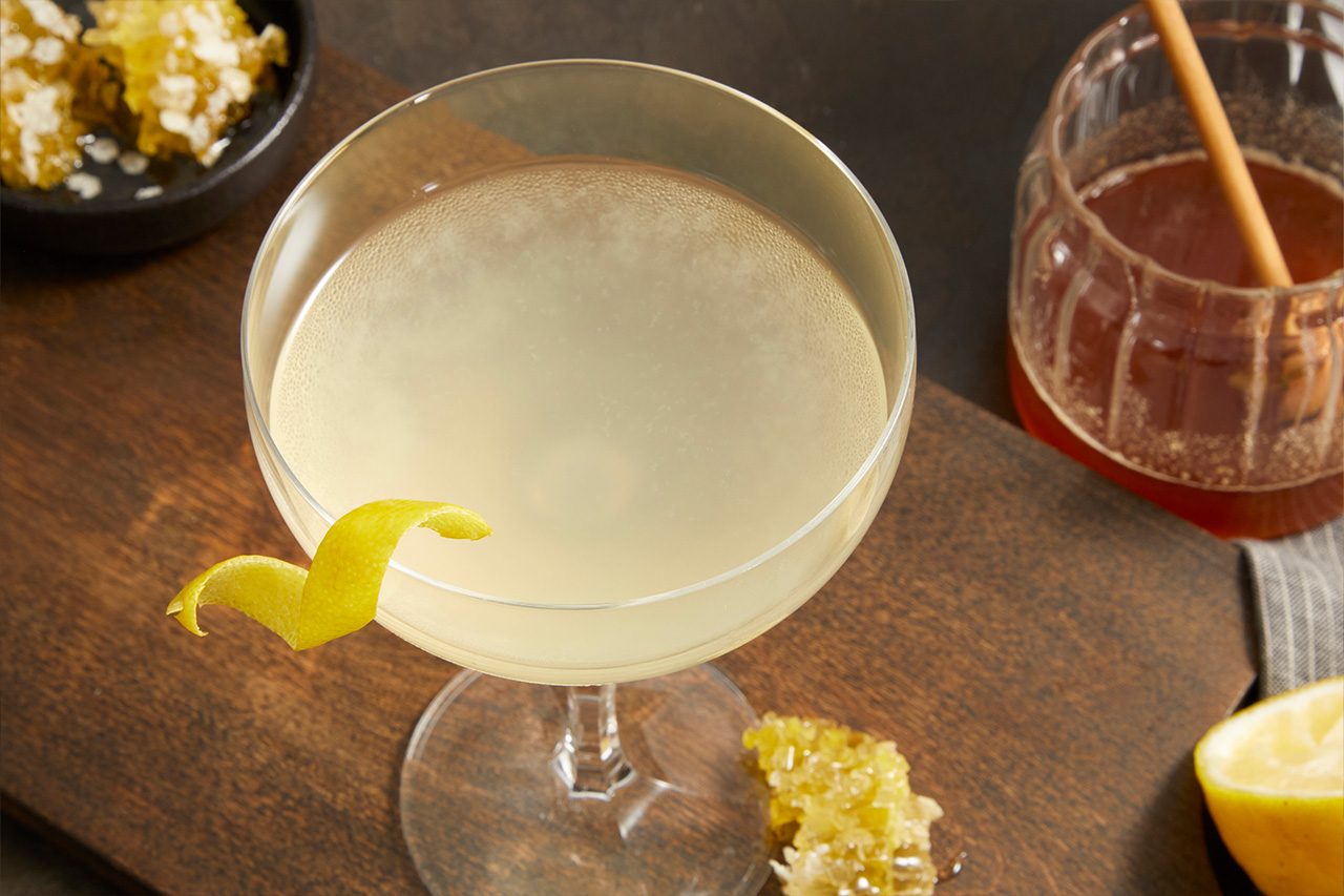 Close-up shot of a Tequila Honey Bee cocktail served in a coupe glass, featuring a pale golden drink garnished with a curled lemon peel. Honey, honeycomb, and a fresh lemon half are arranged nearby on a dark wooden surface.