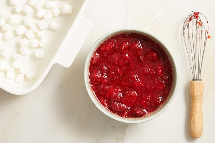 A bowl of red berry mixture sits next to a whisk with a wooden handle and a white dish filled with mini marshmallows, all on a light-colored countertop.