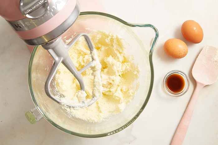 A stand mixer creaming butter and sugar in a glass bowl, with two eggs, vanilla extract, and a pink spatula nearby on a white countertop.