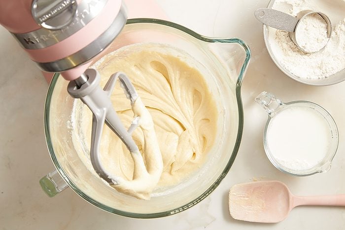 A hand holds a stand mixer, blending creamy batter in a glass bowl. Nearby, there is a cup of milk, a bowl of flour with a sifter, and a pink spatula on a light countertop.