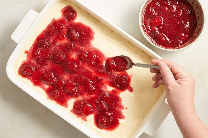 A hand spreads strawberry topping over cake batter in a white baking dish with a spoon. A bowl of strawberry topping sits nearby.